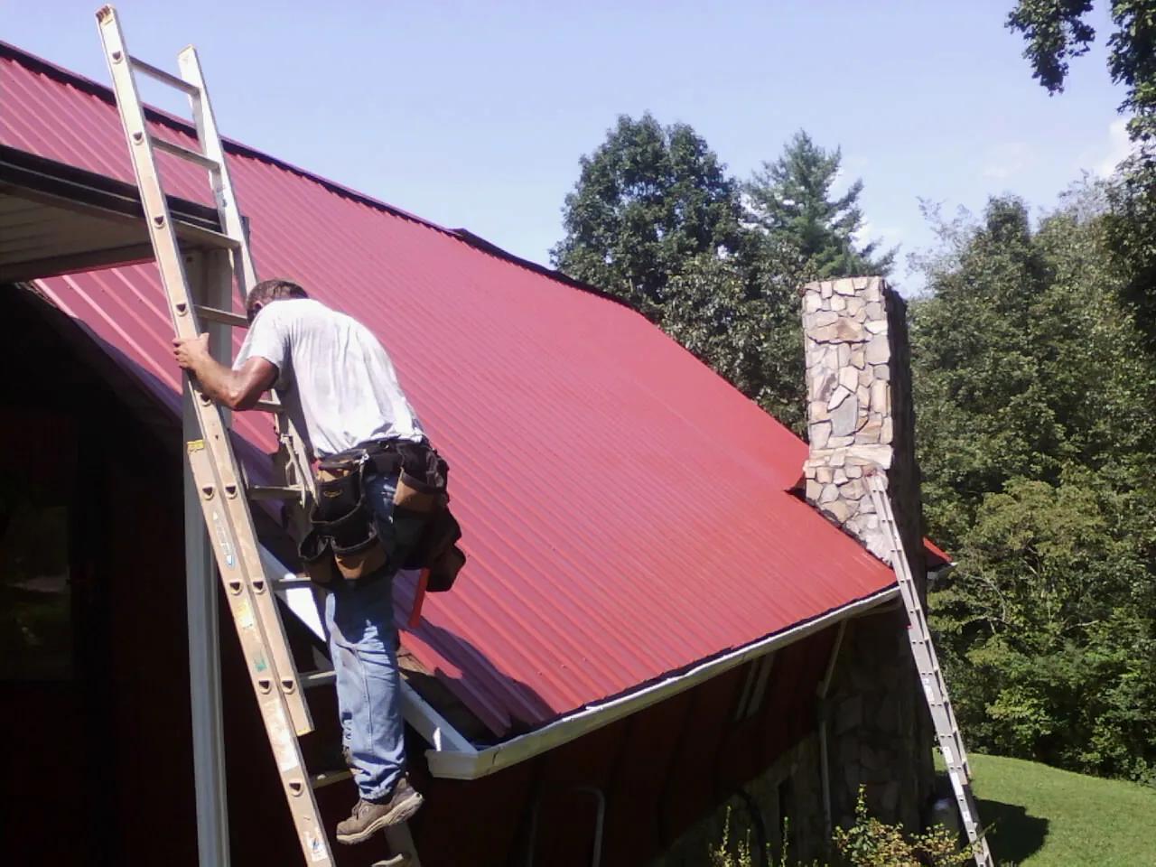 Roofer on a ladder beside a red metal roof and stone chimney — Soffit & Fascia Repair by Roofing Brothers in Manteca